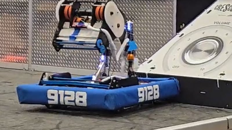 The 9128 robot beside the amp on the Crescendo field, blue bumpers lettered 9128 on two sides, its shooter raised above the chassis.